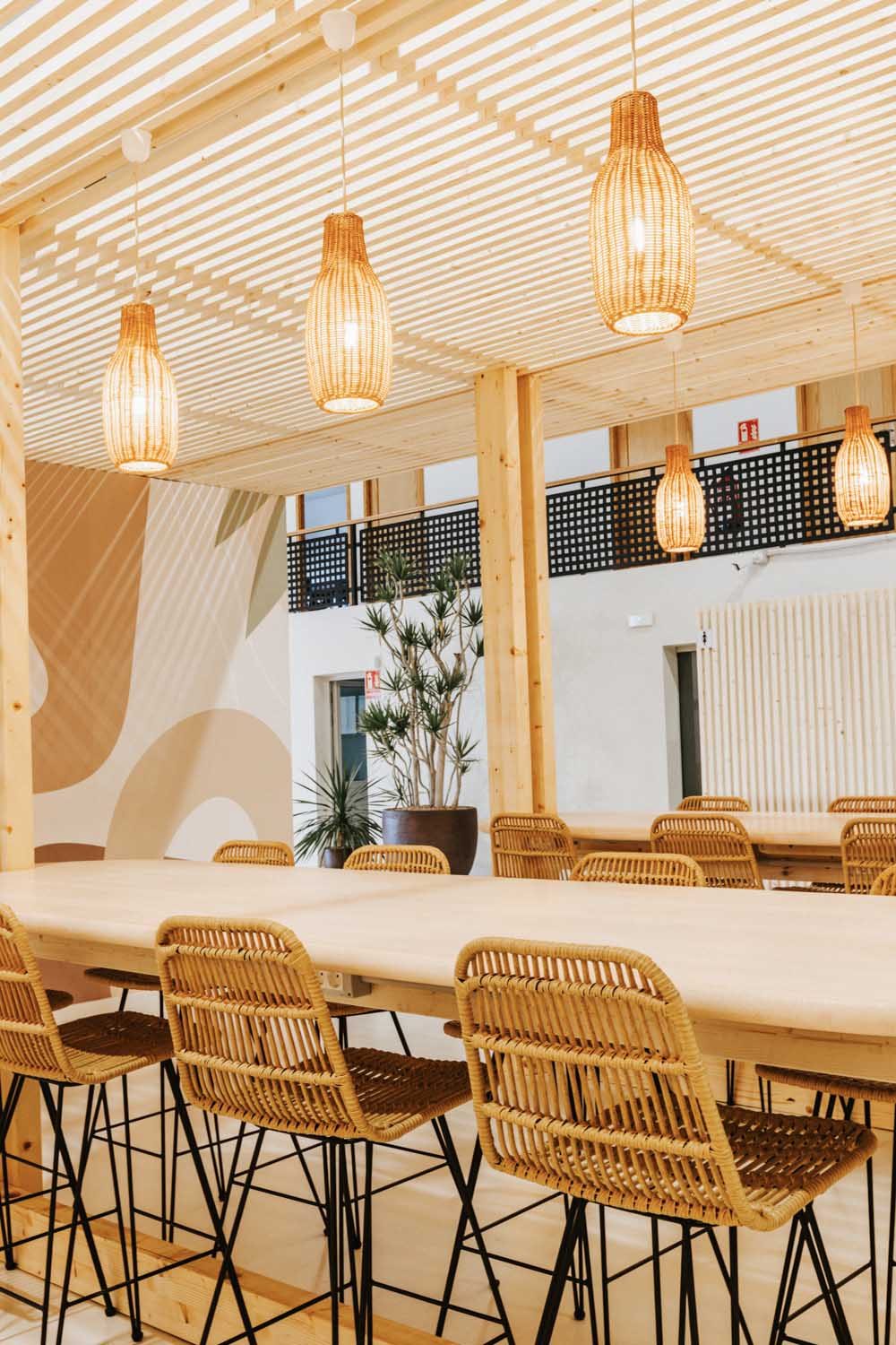 Tall wood-slat pergola ceiling over the communal dining table with rattan pendant lights and potted plants